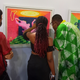 An artist at Art X Lagos 2021 explains her work to two guests on her left and right. The person on the right is looking closely at the painting on the wall.